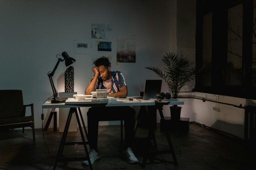 A Black man at his desk struggling to be productive (engaging in busyness without productivity) late at night